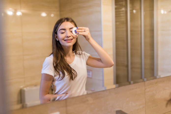 Beautiful young woman cleaning her skin with a cotton pad, looking at the mirror at home bathroom. Beauty, skin care concept, lifestyle Zalety stosowania płynu micelarnego na co dzień